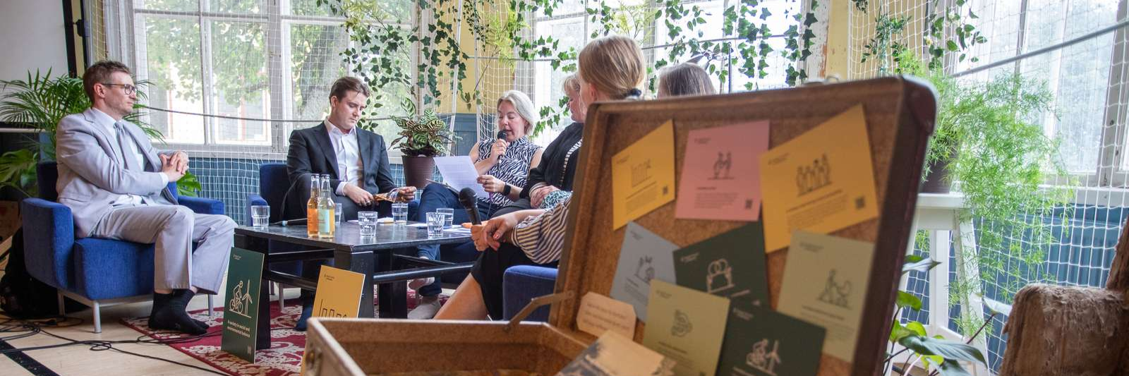 Unga människor sitter i en soffa och diskuterar. I förgrunden en kappsäck. 