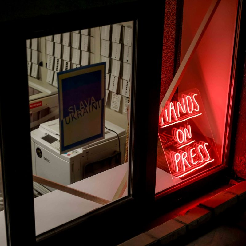 A light sign in a window with the text "Hands on Press"